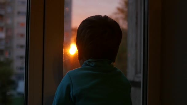 een kind staat op de vensterbank en keek uit het raam in de zon. Zonsondergang - Video