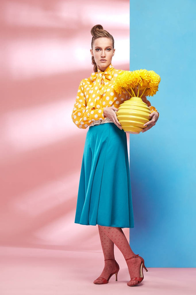 Pretty young sexy woman model wearing yellow blouse with white polka-dot, blue skirt and pink tights in pin-up style, holding vase with yellow flowers and posing in studio with blue and pink background  - Fotó, kép