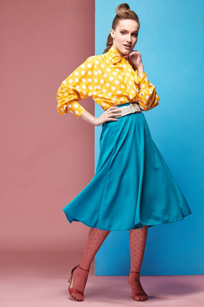 Pretty young sexy serious woman model wearing yellow blouse with white polka-dot, blue skirt and pink tights in pin-up style, posing in studio with blue and pink background  - Fotografie, Obrázek