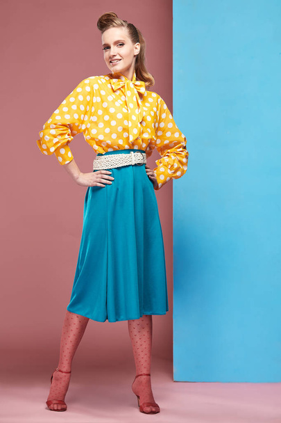 Attractive young sexy smiling woman model wearing yellow blouse with white polka-dot, blue skirt and pink tights in pin-up style, posing in studio with blue and pink background  - Фото, изображение