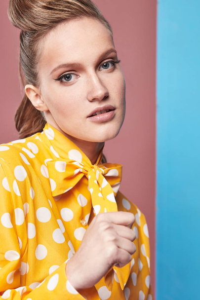Closeup portrait of pretty young sexy woman model wearing yellow blouse with white polka-dot, posing in studio with blue and pink background  - 写真・画像