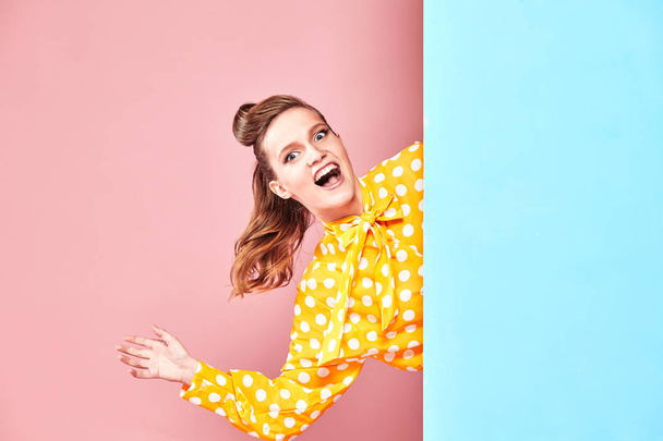Portrait of pretty young woman model wearing yellow blouse with white polka-dot, blue skirt in pin-up style, posing and looking happy in studio with blue and pink background  - Zdjęcie, obraz