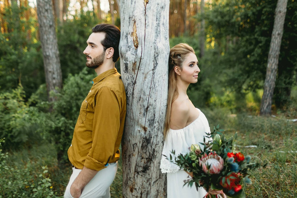 Bride and groom lean on the tree from different sides. Newlyweds are walking in the forest. Artwork - Fotó, kép