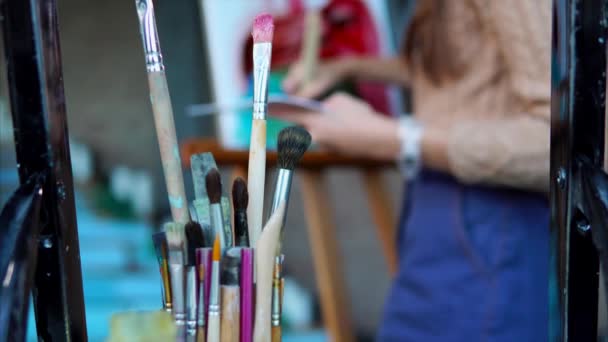 Close up shot of dirty brushes, in the background a woman paints a picture - Footage, Video
