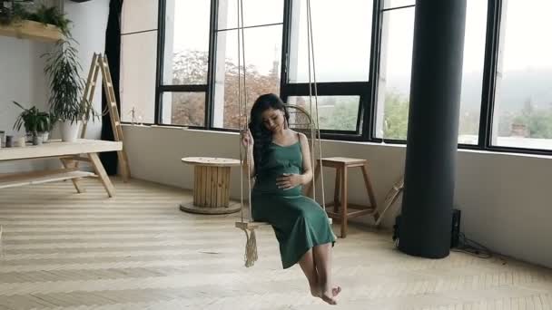 Young pregnant girl swinging on a wooden swing. A young girl in a long green dress on a swing in the studio. - Video, Çekim