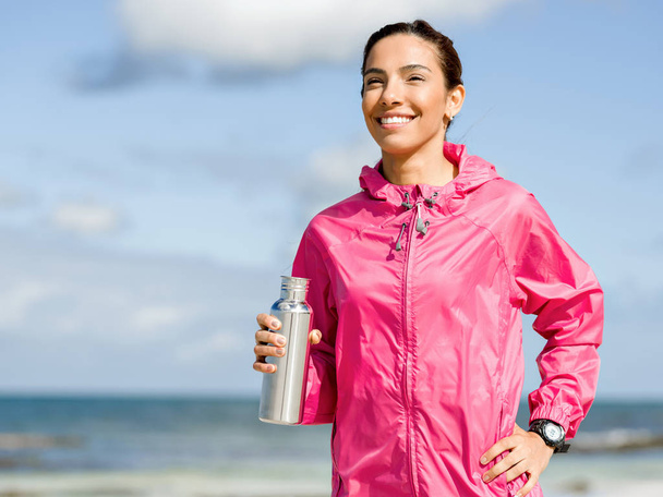 Beautiful girl in sport clothes drinking water after workout on the beach - Foto, Bild