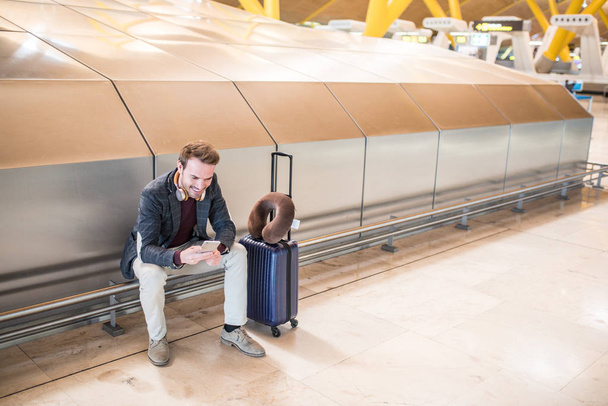 Joven esperando escuchando música y usando un teléfono móvil en el aeropuerto con una maleta
 - Foto, Imagen