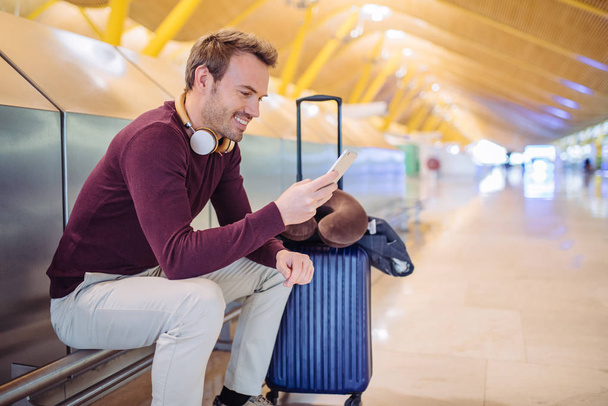Młody człowiek czeka słuchania muzyki i przy użyciu telefonu komórkowego na lotnisku z walizką - Zdjęcie, obraz