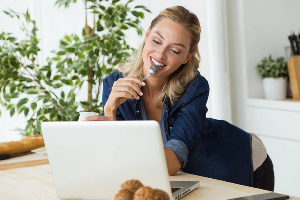Beautiful young woman using her laptop and eating yogurt at home - Foto, Bild