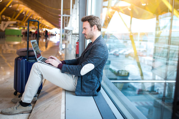 ο άνθρωπος που κάθεται στο αεροδρόμιο χρησιμοποιώντας φορητό υπολογιστή και κινητό τηλέφωνο δίπλα στο παράθυρο. - Φωτογραφία, εικόνα