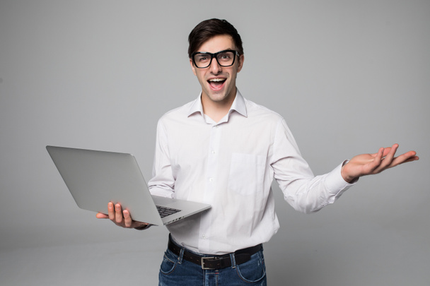 Guapo joven hombre de negocios en camisa y gafas está utilizando un ordenador portátil, mirando a la cámara y sonriendo, sobre fondo gris
 - Foto, Imagen