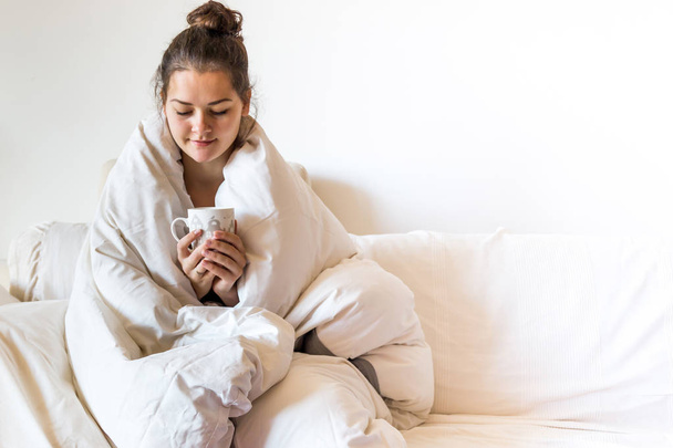 Όμορφη νεαρή γυναίκα που καλύπτεται από τη κουβέρτα είναι κάθεται στο κρεβάτι με καυτό τσάι - Φωτογραφία, εικόνα