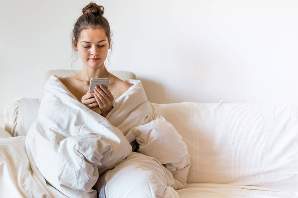 Bella giovane donna coperta da coperta è seduta sul letto con smart phone
 - Foto, immagini