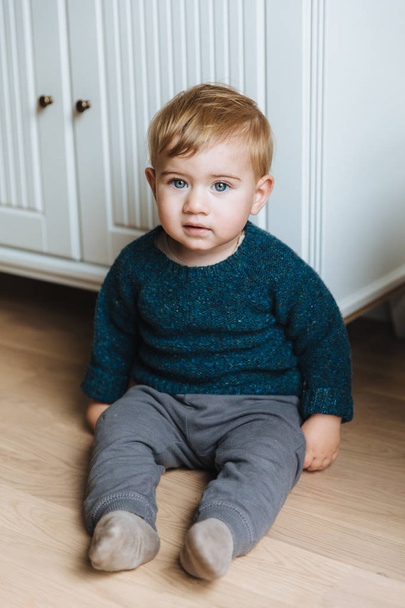 Children concept. Little small infant with appealing blue eyes, plump cheeks and blonde hair sits on floor, teacher to go by himself, wears sweater and trousers. Small child with serious expression - Foto, Imagen