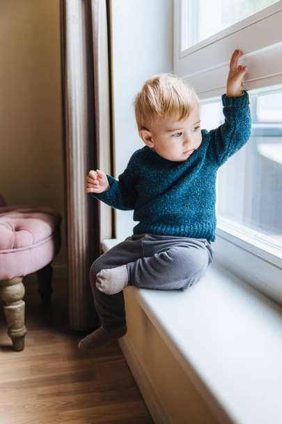 Naughty small kid with blonde hair and blue eyes, sits on window sill, looks out of window, notices something in yard. Curious cute child plays alone, looks outside with attentive expression - Photo, Image