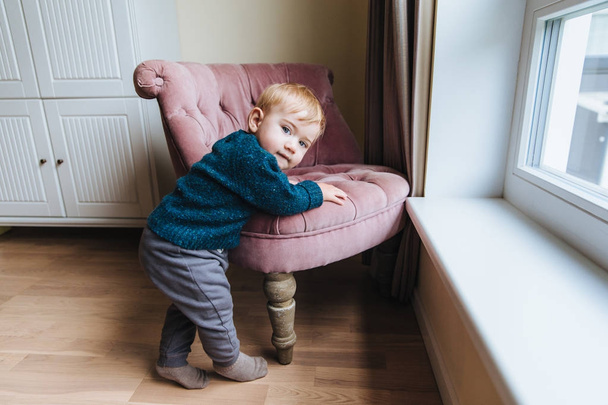 Küçük sarışın çocuk koltuk eğilir, evde tek başına oynuyor. Küçük bebek gitmek öğretir. Çok güzel çocuk yakın sandalyede, yaramaz hileler oynayacak duruyor. Chilren, çocukluk ve yaşam tarzı kavramı - Fotoğraf, Görsel