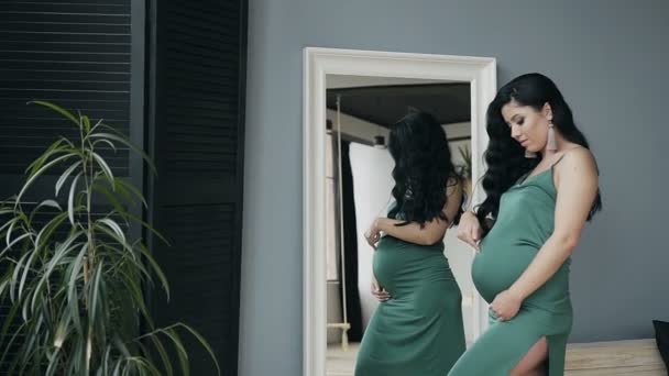 Beautiful pregnant woman standing near a mirror in an evening green dress with beautiful makeup and hairstyle - Materiał filmowy, wideo
