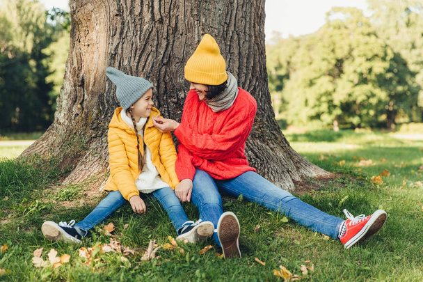 Little daughter and her mother have fun together, dressed warm, sit near big tree on green grass, look at each other with love. Affectionate mom and small female kid have good mood, spend weekends - Photo, Image