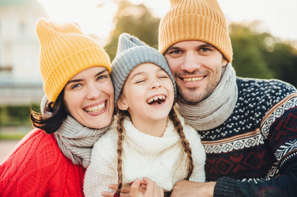 Horizontal portrait of smiling happy woman and man have good time with their daughter, wear knitted hats, warm sweaters and scarfs, going in park to have walk together. Spending time in family circle - Photo, Image