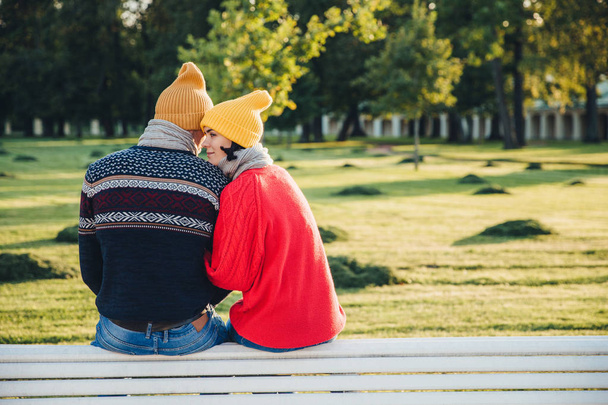 Lovely couple sit on bench together, wear warm clothes and knitted hats, embrace each other, express love and good relationship, enjoy beautiful landscapes and fresh air in park. Relationship concept - Photo, Image