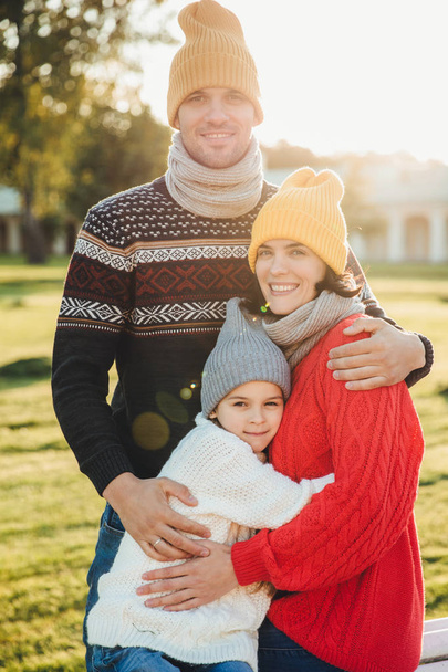 Vertical portrait of friendly family couple stand together, embrace each other, have good relationships, enjoy sunny weather. Handsome unshaven man hugs his wife and daughter, pose outdoors - Photo, Image