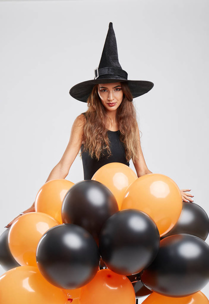 Sexy girl in a witch costume with black and orange balloons on a gray background. Halloween witch concept. - Фото, зображення