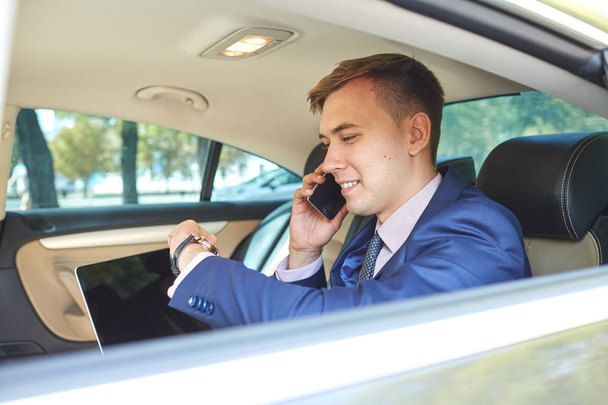 Businessman looking at the clock in his car with a laptop talking on the phone - Photo, Image