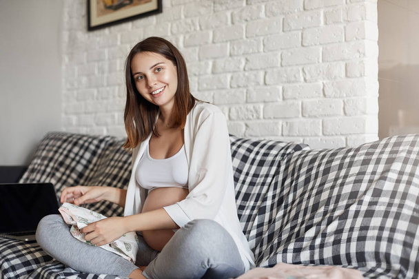Portrait of young pregnant female preparing clothes for her newborn baby looking at camera smiling sitting in a cozy sofa at home. Pregnancy and expectations concept. - Photo, Image