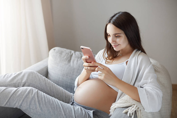 Young pregnant lady using smart phone to get all the status updates of her friends. Reading about meds to fight nausea and stay happy away from depression. - Фото, изображение