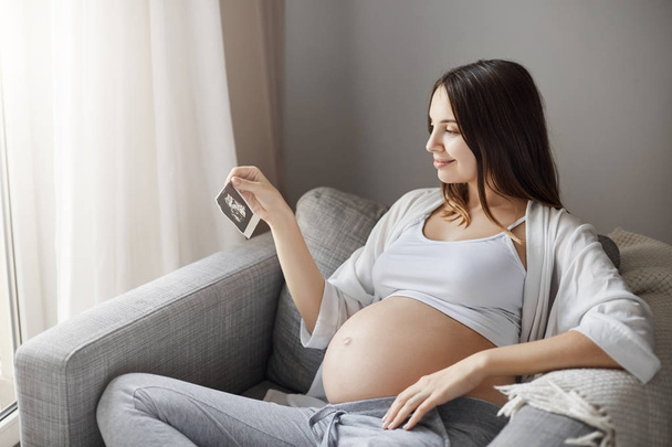 Zwangere dame op zoek naar een baby echografie foto probeert te achterhalen is het een jongen of een meisje. Home verblijf in haar gezellige Bank. - Foto, afbeelding