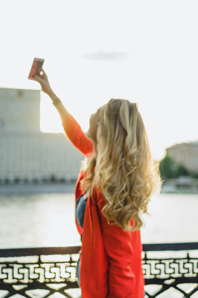 back of young blonde woman in red jacket with phone outdoor - Foto, imagen