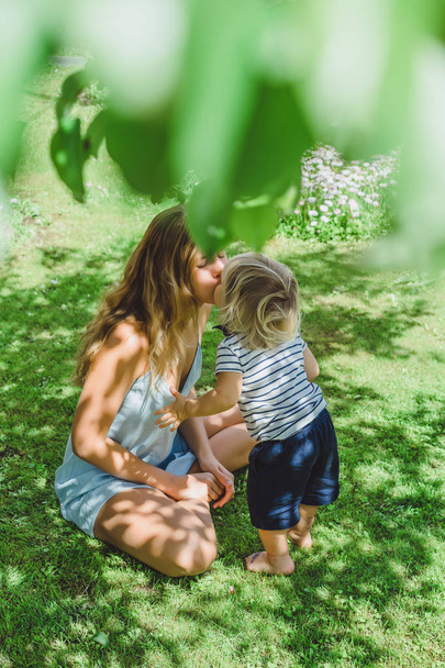 young mother kissing cute blonde boy on green grass outdoor at summer - Фото, зображення