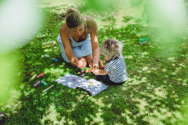 cute blonde boy drawing with young mother on green grass outdoor - Foto, immagini
