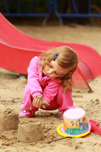Jeden rok starý roztomilá kudrnatá dívka v barvě růžové sportovní hrát a dělat hrady z písku na hřišti - Fotografie, Obrázek