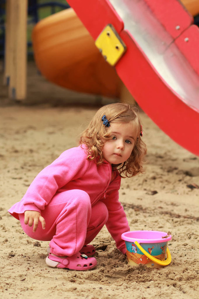 Um ano de idade bonito menina encaracolado em rosa terno de esportes jogando e fazendo castelos de areia no parque infantil
 - Foto, Imagem