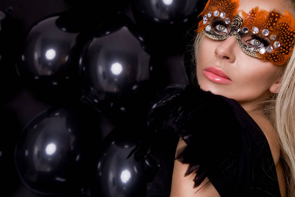 Mooie blonde sensuele vrouw met carnaval masker, staande op een achtergrond van zwarte ballonnen en sexy in de lens kijkt - Foto, afbeelding