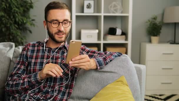 Handsome caucasian man in checked casual shirt and glasses is having a great time at home, sitting on the couch and videochatting with friends with earbuds on his smartphone smiling and waving to the - Séquence, vidéo