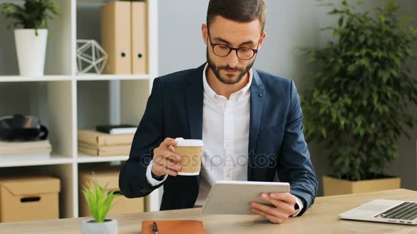 Busy young man in the office sipping coffee and looking at his tablet device on the office background. Indoor shot. - Кадри, відео