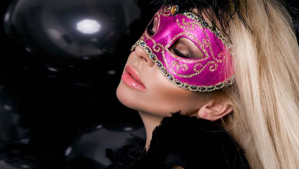 Mulher loira sensual bonita com máscara de carnaval, de pé sobre um fundo de balões pretos e sexy olha para a lente
 - Foto, Imagem