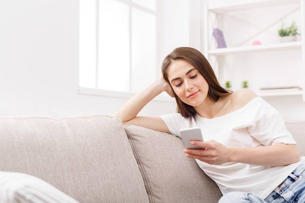 Young woman using smartphone at home on the couch. Dark-haired girl in casual texting online - Foto, Imagem