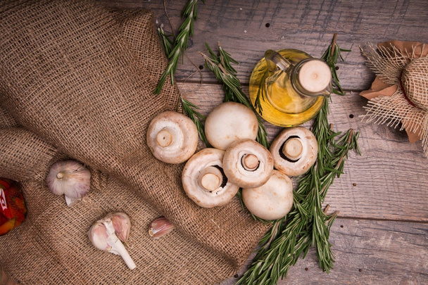 A view from above of a heap of fresh white mushrooms from a forest on a wooden background. A vase full of sunflowers oil, green rosemary and spicy garlic on an old brown bag on the table. - Valokuva, kuva
