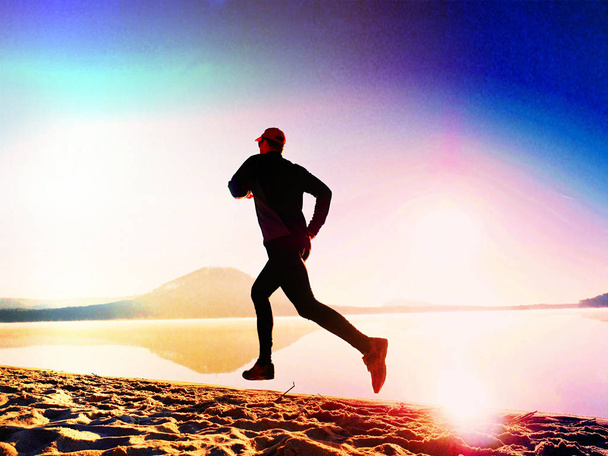Tall fit man running along the sea shore in morning. Healthy male jogging on lake beach. - Photo, Image