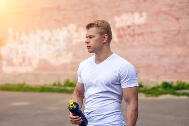 An athlete in nature, after training, in his hand a bottle of water, a shaker with a protein. The coach controls the training in the city. - Valokuva, kuva