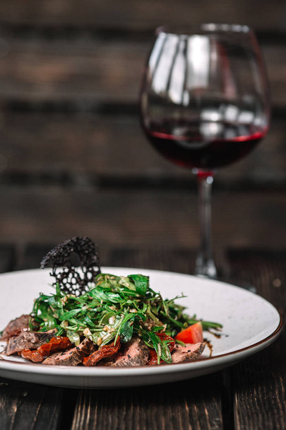 Glass of wine and salad with warm veal, sun dried tomatoes and arugula on dark wooden background - Photo, Image