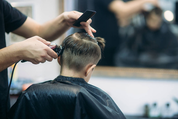 Pequeño chico consiguiendo corte de pelo por peluquero mientras está sentado en silla en la barbería
. - Foto, Imagen