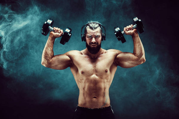 Человек тренирует мышцы с гантелями в студии на темном фоне с дымом
. - Фото, изображение