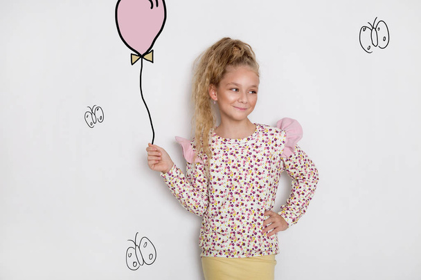 atemberaubend schöne kleine Mädchen mit langen blonden Haaren steht auf einem weißen Hintergrund und hält einen Luftballon und Blumen in seiner Hand - Foto, Bild