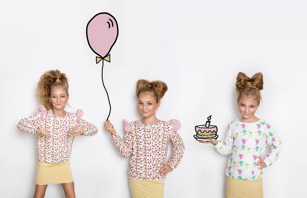 drei atemberaubende kleine Mädchen mit langen blonden Haaren stehen auf weißem Hintergrund und eines von ihnen hält einen Luftballon, das zweite und dritte tanzen mit einer Geburtstagstorte - Foto, Bild