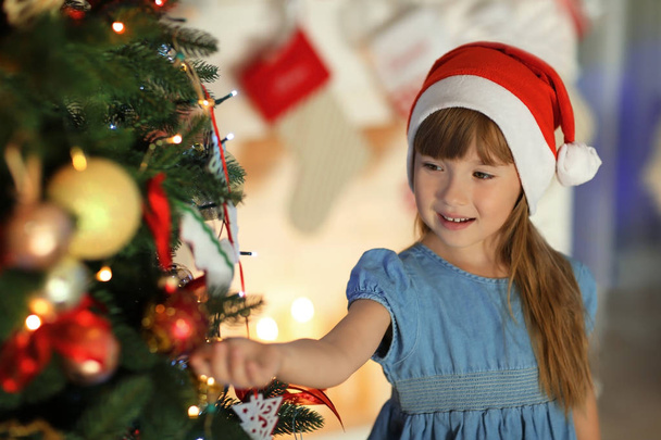 Cute little girl in Santa hat decorating Christmas tree at home - Photo, Image