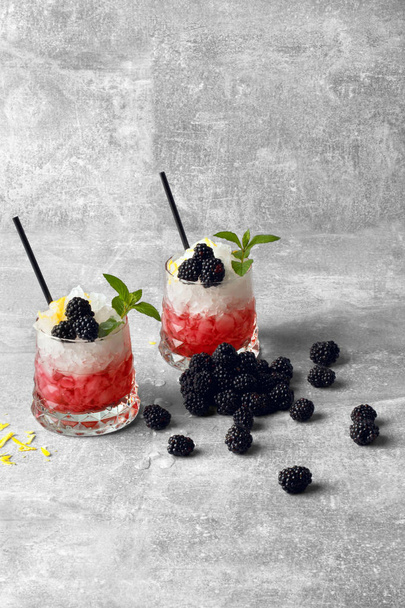A gray table with sappy blackberries, two glasses of red fresh juice with pieces of ice and black drinking straws, yellow lemon peel on a light gray background. Refreshing drinks. - Foto, Imagen